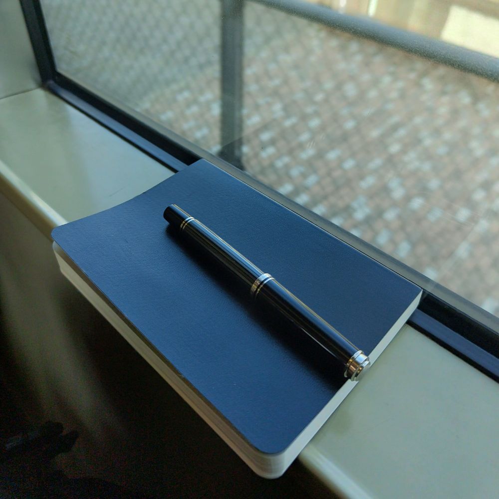 黒い小さなノート、黒い万年筆が電車の窓の横に置かれている写真